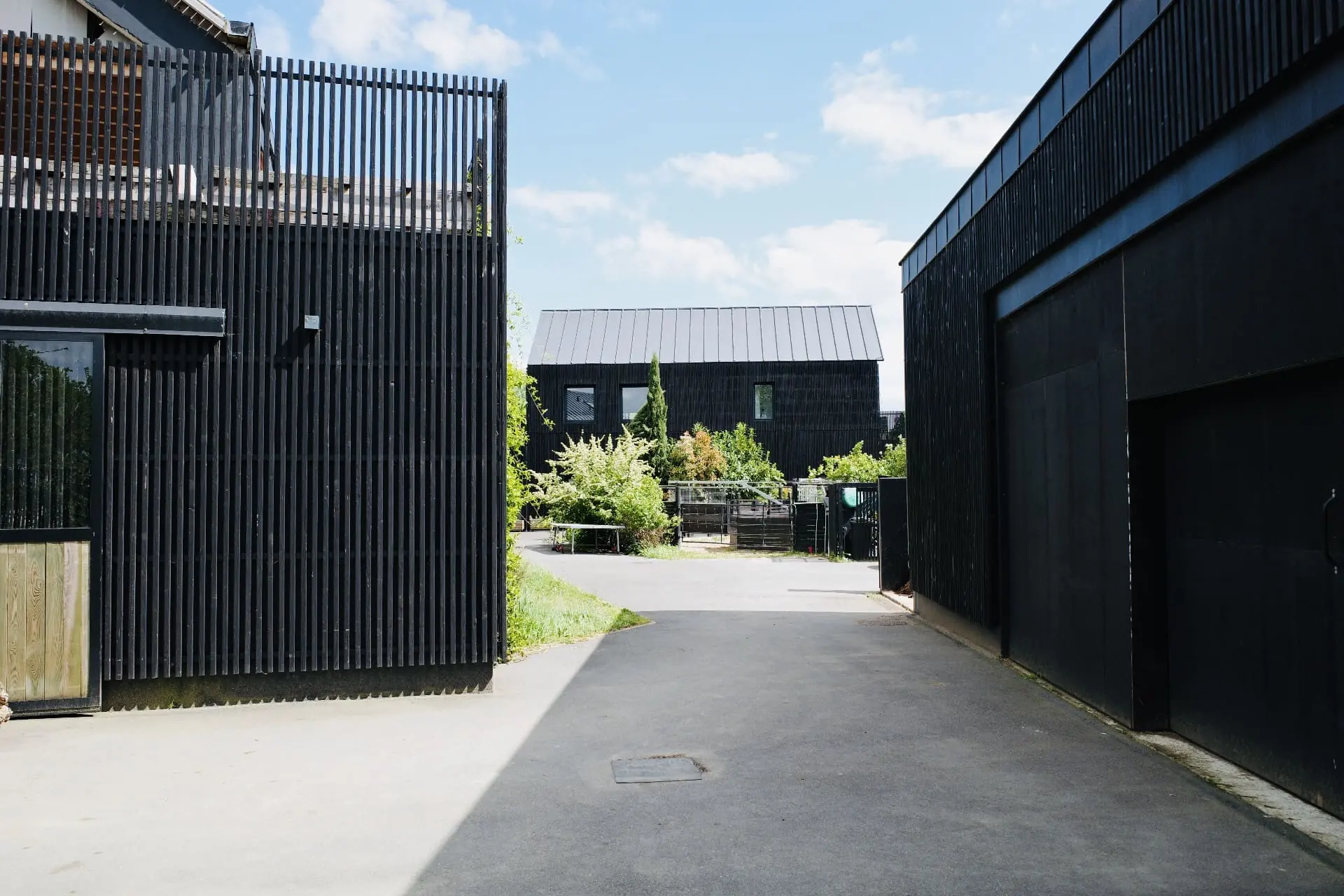 Passage entre deux bâtiments en bardage noir ouvrant sur la cour centrale, la maison à pignon et toiture zinc se détachant au fond.