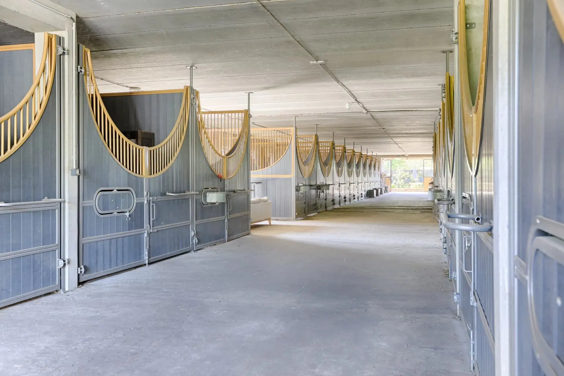 Allée centrale de l'écurie, boxes en enfilade de part et d'autre avec grilles bois et métal, ouverture éclairée au fond.
