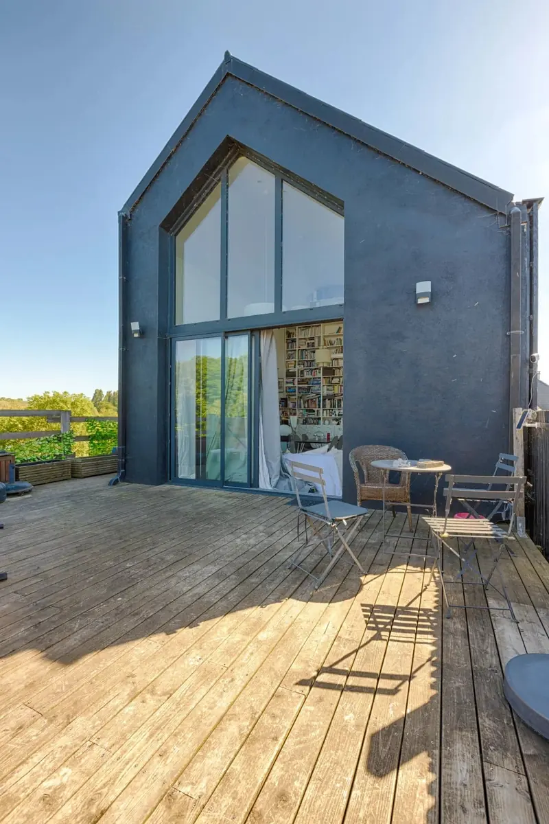 Terrasse en bois de l'étage devant une grande baie vitrée toute hauteur, ouverte sur une pièce tapissée de livres.